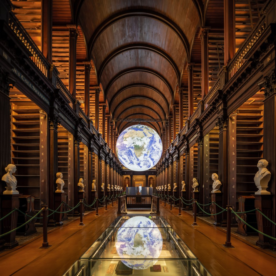 A large globe displayed in the Long Room of the Old Library, with empty bookshelves and marble busts in the background.